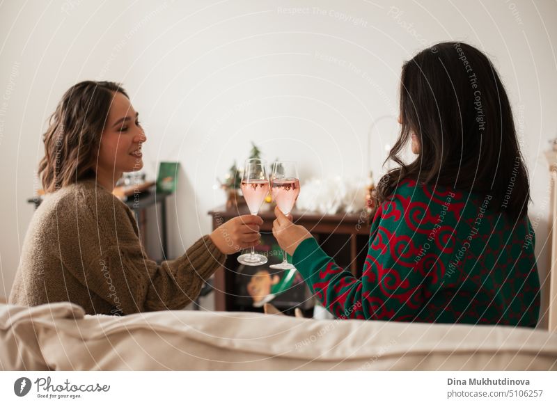 Zwei Frauen stoßen mit Sektgläsern auf einer Party an, trinken, um einen Erfolg oder einen Feiertag zu feiern. St. Valentines oder Galentine's Day. Freunde feiern Urlaub Saison sitzen auf einer Couch zu Hause Wohnung und trinken, glücklich zu sein, lächelnd und lachend.