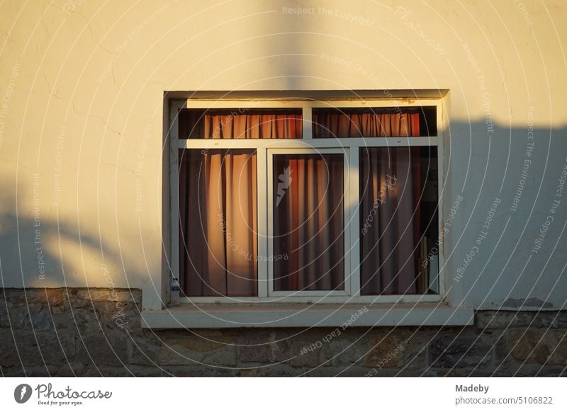 Fassade in Beige und Naturfarben im Licht der Abendsonne mit weißem Fenster mit rotem Vorhang in Cunda bei Ayvalik am Ägäischen Meer in der Provinz Balikesir in der Türkei