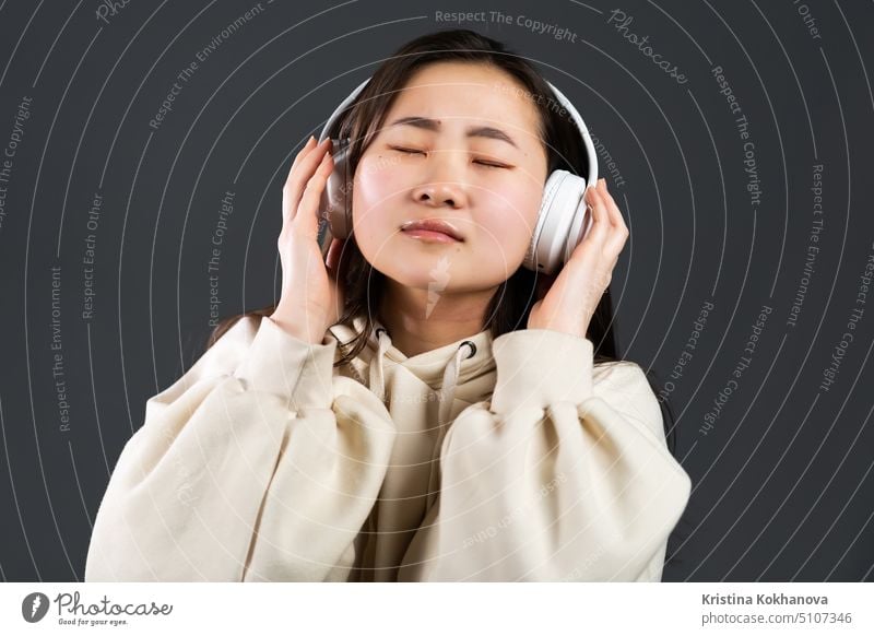 Junge schöne asiatische Frau mit Kopfhörern in moderner Kleidung genießt und tanzt auf grauem Hintergrund. Radio, Melodie, Technologie-Konzept. Licht Glamour