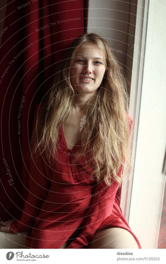Nahes Portrait einer jungen, blonden Frau, die lächelnd in rotem Kleid am Fenster sitzt junge Frau langhaarig Freude schlank freundlich angenehm schön Porträt