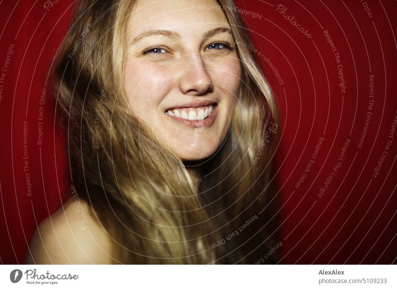 Nahes Portrait einer jungen, blonden Frau, die lächelnd vor einem roten Hintergrund steht junge Frau langhaarig Freude schlank freundlich angenehm schön Porträt