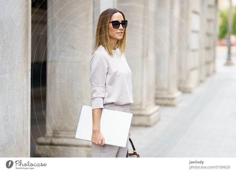 Geschäftsfrau mit Sonnenbrille auf der Straße stehend Laptop Großstadt selbstbewusst Arbeitsweg urban Stil Outfit Apparatur Handtasche freiberuflich Gebäude