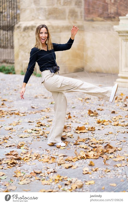 Fröhliche junge Frau, die beim Spazierengehen im Park gegen heruntergefallene Blätter tritt Spaziergang Lächeln Feiertag Herbst Tourist heiter Urlaub genießen
