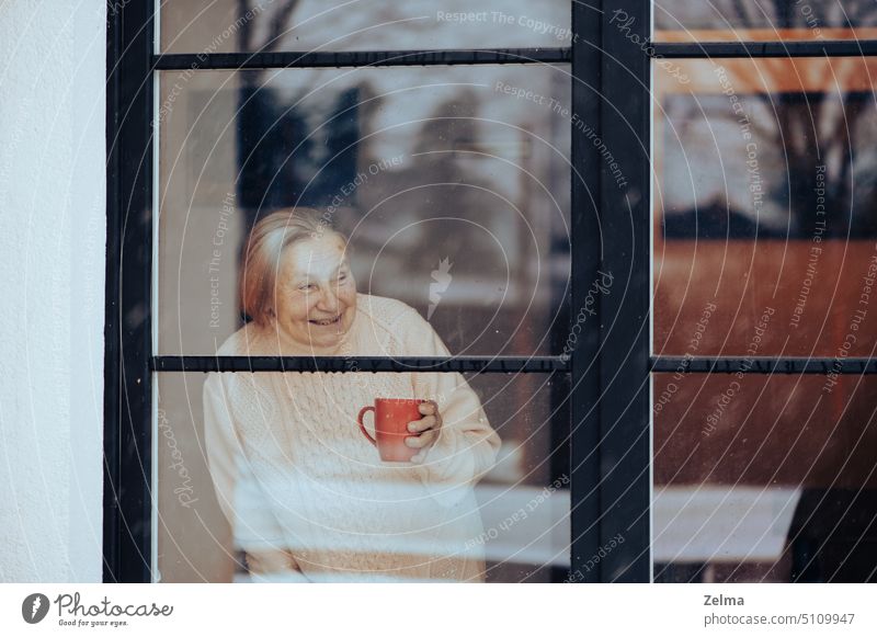 lächelnde alte Frau mit einer Tasse Kaffee schaut durch ein schmutziges Fensterglas Senior älter Blick dreckig Glas Lächeln Gesicht Pflegeheim Haus