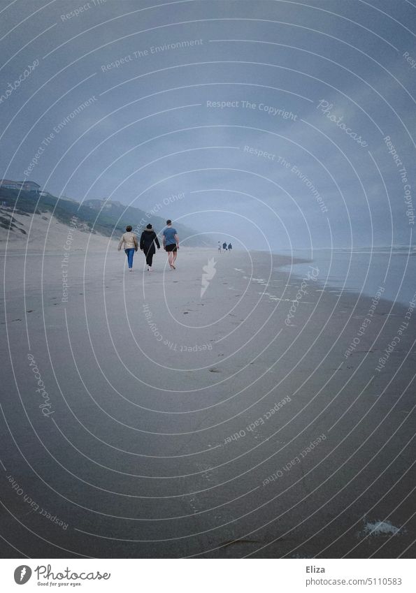 Menschen spazieren bei bewölktem Wetter am Strand entlang grau düster Meer trist Küste Wolken drei Spaziergang Strandspaziergang am Meer Erholung Urlaub trüb