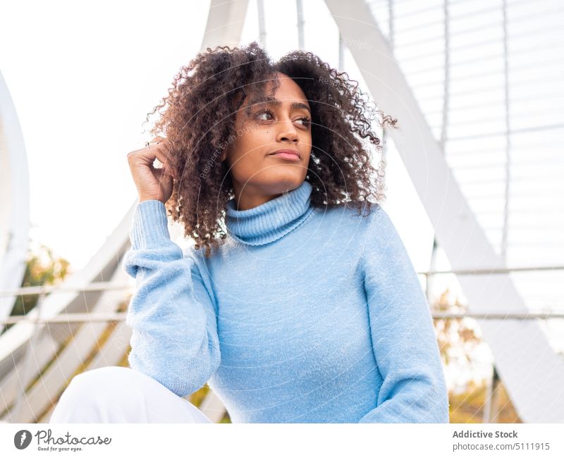 Ethnische Frau mit Afro-Haar sitzt im Park verträumt Haare berühren nachdenken Bank positiv lässig Herbst modern ruhen jung hispanisch besinnlich nachdenklich