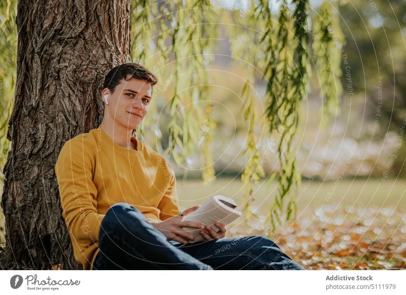 Glücklicher Mann sitzt unter einem Baum und liest ein Buch benutzend Kopfhörer Park Natur Weide Herbst Apparatur männlich Gerät Lächeln Laubwerk zuhören Blatt