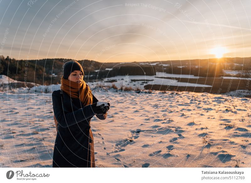 Eine junge Frau in Mantel und Hut steht bei Sonnenuntergang auf einem schneebedeckten Feld und hält eine Handvoll Schnee in ihren Händen stehen Halt Hand.