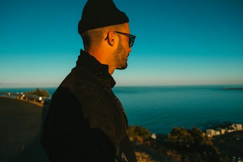 Mann mit Sonnenbrille und Mütze betrachtet das Meer von einem Aussichtspunkt Mensch blau seitlich Seitenansicht nachdenklich Erwachsen Hipster blauer Himmel