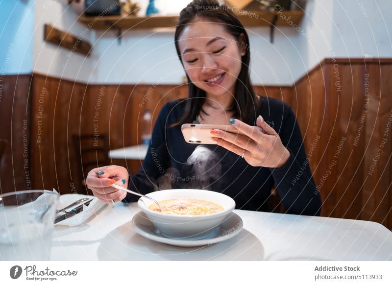 Fröhliche junge Asiatin, die eine Suppe im Restaurant fotografiert Frau fotografieren Lächeln Smartphone Abendessen lecker Ramen Lebensmittel Freude Mobile