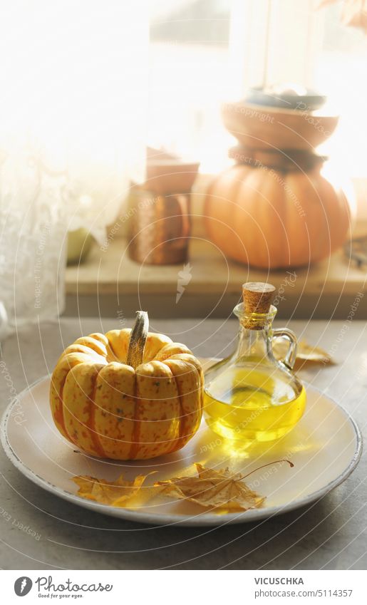 Kleiner Kürbis, Speiseöl in Glaskaraffe und gelbes Herbstblatt auf Teller am Fensterhintergrund mit natürlichem Licht. Kochen zu Hause mit Herbstgemüse der Saison. Vorderansicht.
