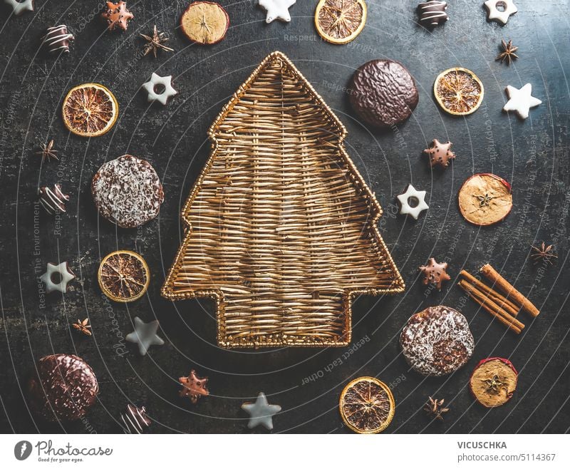 Weihnachtsbaumförmiges Holztablett auf dunklem Betontisch mit Lebkuchen, Keksen, getrockneten Orangenscheiben und Zimtstangen. Leckere saisonale Wintersüßigkeiten und Aromen. Ansicht von oben.