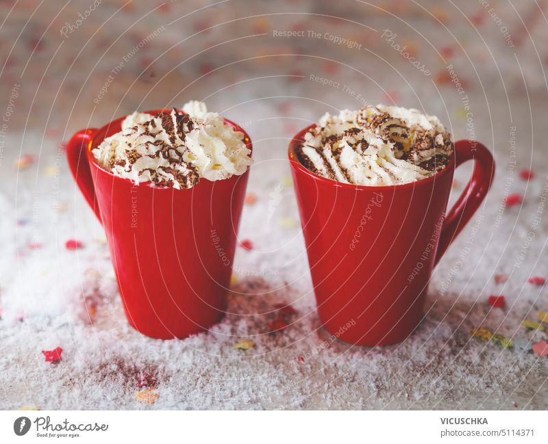 Zwei rote Becher mit heißer Schokolade und Schlagsahne auf dem Tisch mit Schnee. Gemütliche warme Getränke zu Hause mit süßer Sahne. Vorderansicht. zwei Kakao