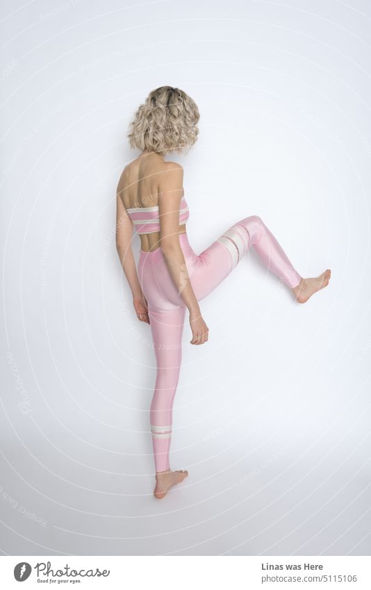 Ein minimalistisches Shooting in einem Studio. Ein Modellversuch mit einem blondgelockten Mädchen. Sie tritt in ihrem rosa glänzenden Sportoutfit gegen eine Wand. Sportmode mit etwas Action.