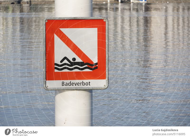Roter quadratischer Wegweiser mit der Aufschrift "Baden verboten" in deutscher Sprache an einem Metallmast befestigt. Platziert im Vierwaldstättersee in der Schweiz. Der See selbst ist im Hintergrund zu sehen. Es ist ein Warnsymbol vor Gefahren.