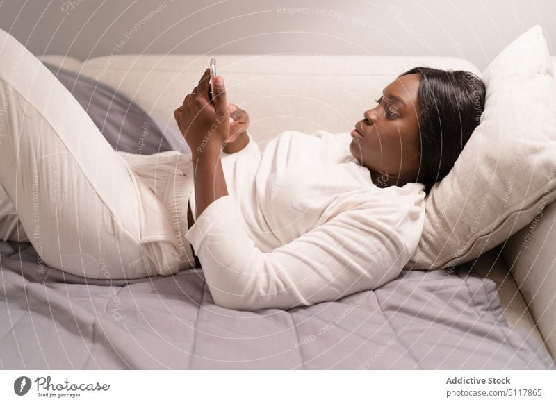 Afroamerikanische Frau benutzt Smartphone auf Sofa benutzend soziale Netzwerke Pyjama ruhen heimwärts Abend Süchtige jung schwarz Afroamerikaner Decke