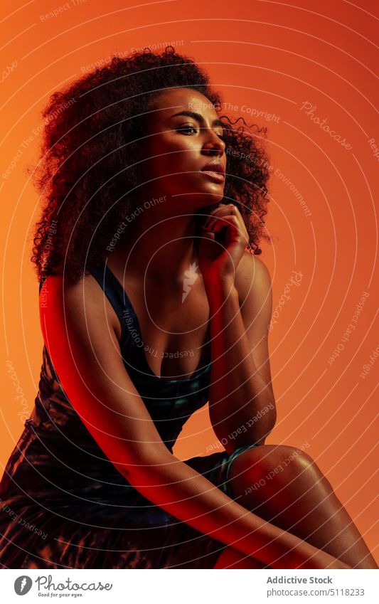 Verführerische ethnische Frau im Kleid sinnlich Verlockung Model Kinn berühren Beine gekreuzt Afro-Look Studioaufnahme Stil feminin Vorschein elegant