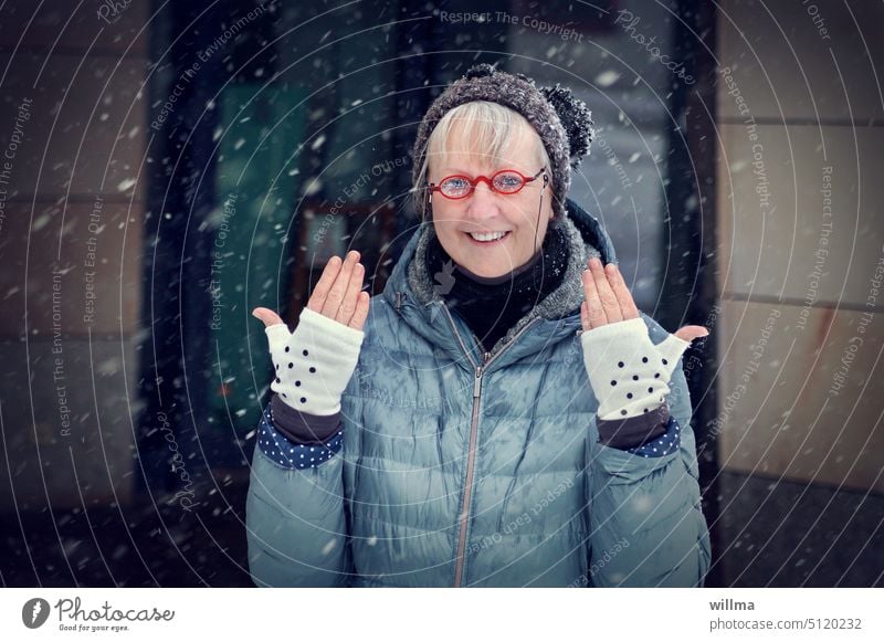 Ältere Frau mit roter Brille und Bommelmütze lacht und zeigt ihre Handschuhe mit Pünktchen Winter fröhlich fingerlose Handschuhe Hände Freude Mütze lachen