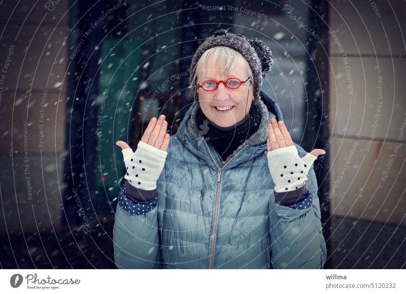 Fröhliche Frau mit roter Brille und Bommelmütze zeigt den Schneeflocken ihre Handschuhe mit Pünktchen Winter fröhlich fingerlose Handschuhe Hände Freude Mütze