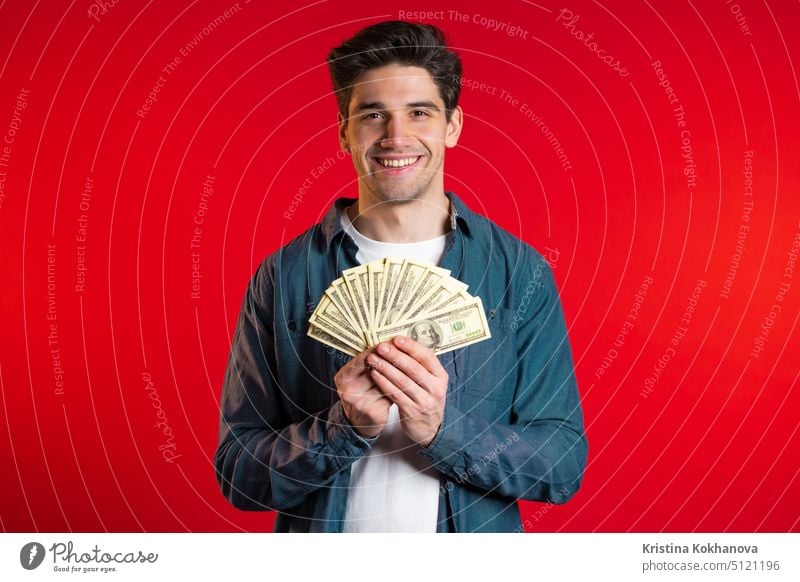 Zufriedener glücklicher aufgeregter Mann zeigt Geld - US-Währung Dollar-Banknoten auf roter Wand. Symbol für Erfolg, Gewinn, Sieg. reich Bargeld Finanzen