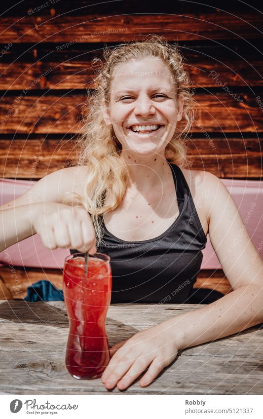 Eine junge blonde Frau mit einem Getränk an einem Holztisch junge Frau lächelnd lächelnde Frau Lachen freundlich Blick in die Kamera Berghütte Hütte Pause Rast