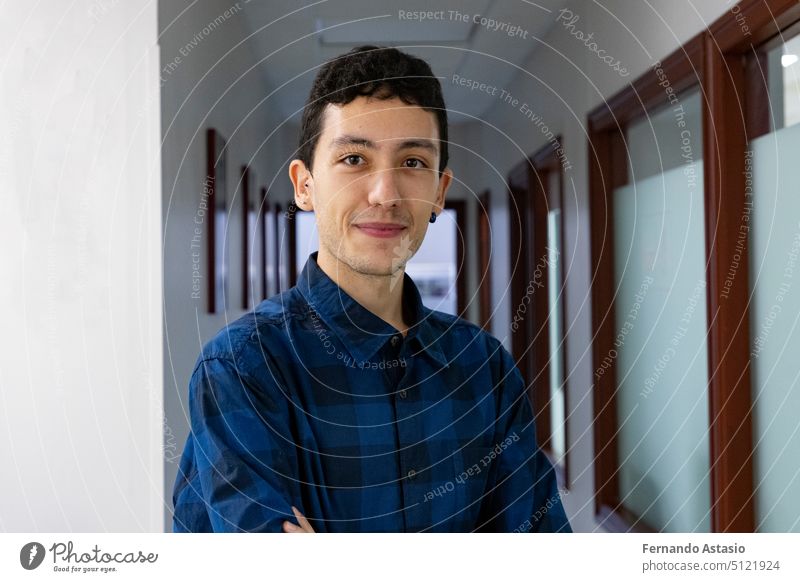 Porträt eines lächelnden jungen kaukasischen Mannes mit gekreuzten Händen in einem langen Korridor, der ein blau-schwarz kariertes Hemd trägt. Porträtfotografie. Modell. Junge mit Ohrringen.
