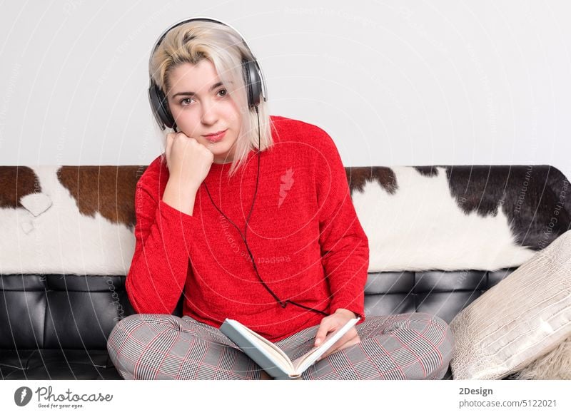 Gelangweilte Frau mit Kopfhörern, die auf einer Ledercouch sitzt und ein Buch hält blond Liege lesen rot Sitzen gelangweilt Barfuß Pullover im Innenbereich jung