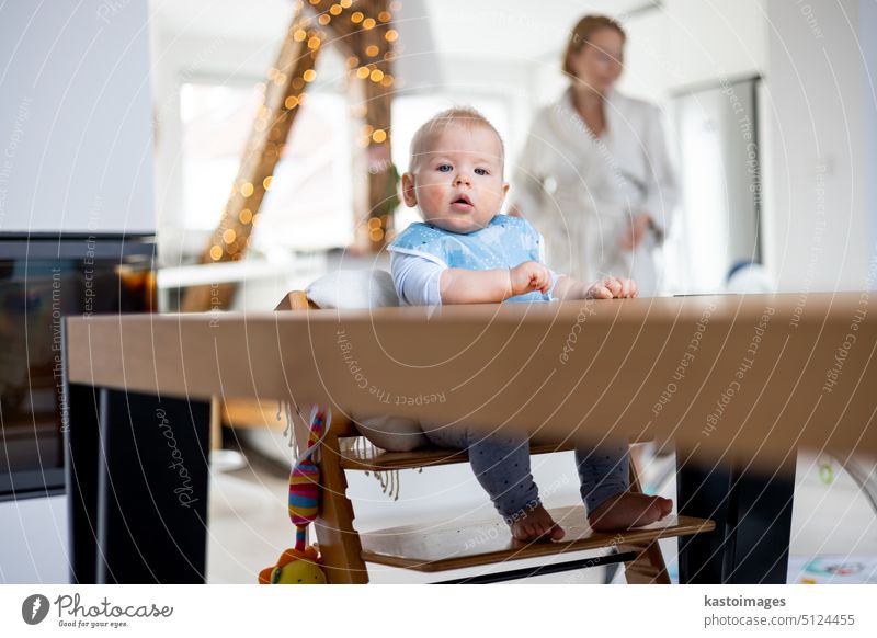 Glückliches Kleinkind, das am Esstisch sitzt und mit seinem Spielzeug in einem traditionellen skandinavischen Designer-Hochstuhl aus Holz in einem modernen, hellen, von seiner Mutter betreuten Haus spielt.