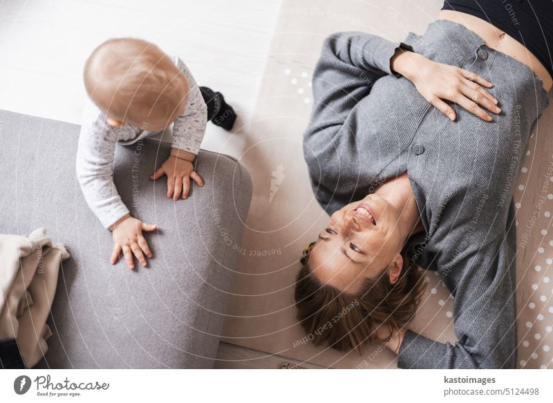 Glückliche Familienmomente. Mutter liegt bequem auf der Kindermatte und spielt mit ihrem kleinen Jungen, der seine ersten Schritte beobachtet und überwacht. Positive menschliche Emotionen, Gefühle, Freude.