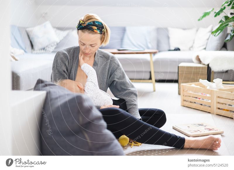 Junge Frau, die ihr Baby stillt, sitzt lässig auf der Spielmatte des Kindes auf dem Wohnzimmerboden zu Hause. Eltern Mutter Stillen Mutterschaft Säugling