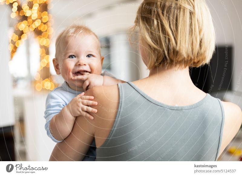Blonde Mutter hält ihr bezauberndes fröhliches kleines Baby im Arm. Nettes Kleinkind Kind in die Kamera schauen und lächelnd zu Hause Junge Säugling Beteiligung