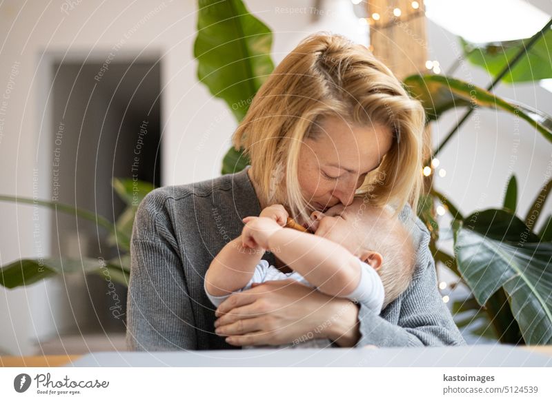 Porträt einer jungen Mutter, die ihr bezauberndes kleines Kind kuschelt und küsst, während sie zu Hause am Tisch sitzt. Sinnesanregungen für die Entwicklung des Babys