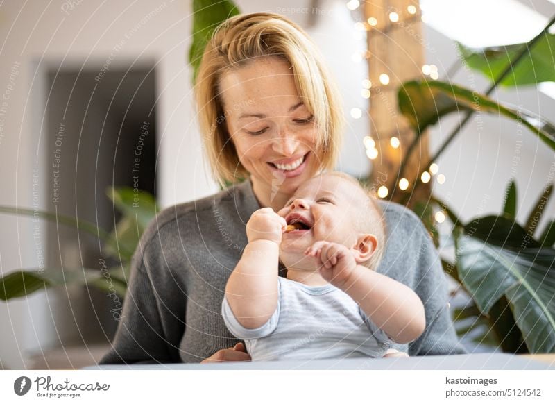 Porträt einer jungen Mutter, die ihr bezauberndes kleines Kind kuschelt, während sie zu Hause am Tisch sitzt. Sinnesanregung für die Entwicklung des Babys.