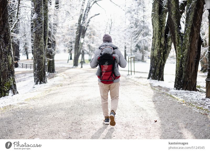 Rückansicht eines sportlichen Vaters, der seinen kleinen Sohn mit Winteroverall und Mütze im Rucksack auf dem Rücken durch einen winterlichen Stadtpark trägt