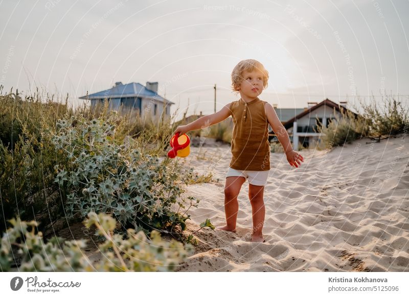 Süßer kleiner Junge gießt Pflanzen mit Gießkanne am Sandstrand. Sommer sonniger Tag. Kleinkind mit buntem Topf. Natürliche ästhetische Porträt des Kindes.