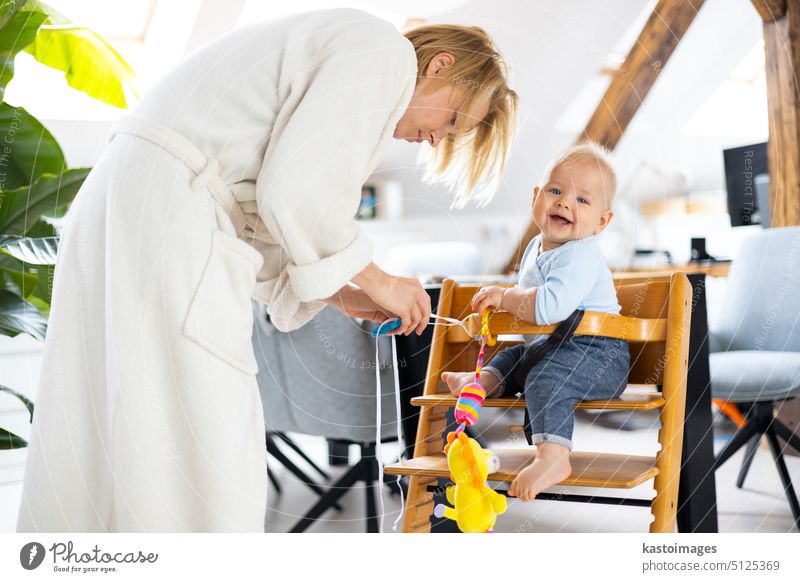 Glückliches Kleinkind sitzt und spielt mit seinem Spielzeug in einem traditionellen skandinavischen Designer-Hochstuhl aus Holz in einem modernen, hellen Haus, das von seiner Mutter betreut wird