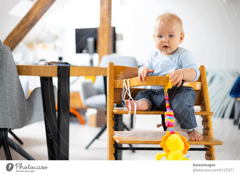 Glückliches Kleinkind sitzt und spielt mit seinem Spielzeug im traditionellen skandinavischen Designer-Hochstuhl aus Holz in einem modernen, hellen Haus. Niedliches Baby.
