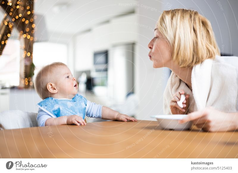 Mutter motiviert ihr Baby Junge Kleinkind Kind während Löffel füttern ihn sitzen in Hochstuhl am Esstisch zu Hause. Baby feste Nahrung Einführung Konzept