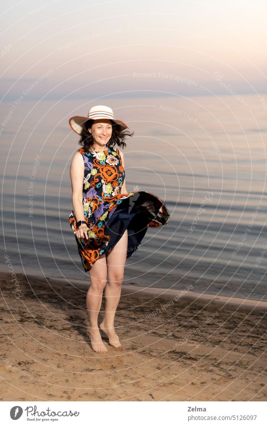 Fröhliche junge Frau in geblümtem Kleid und Sommerhut tanzt fröhlich am Meeresufer Glück Mädchen rennen Tanzen Strand Sonnenuntergang Hut Lächeln springend Sand