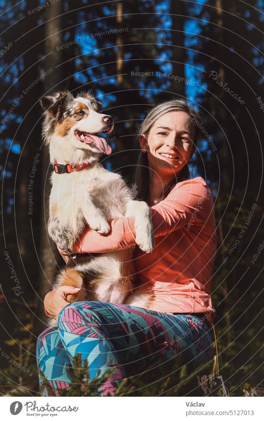 Unverblümtes Porträt einer jungen Sportlerin mit ihrem Lauf- und Wanderpartner, einem Australian Shepherd Hund. Realistisches Lächeln von Frau und Hund. Glückliches Paar. Blue Merle und Wanderer