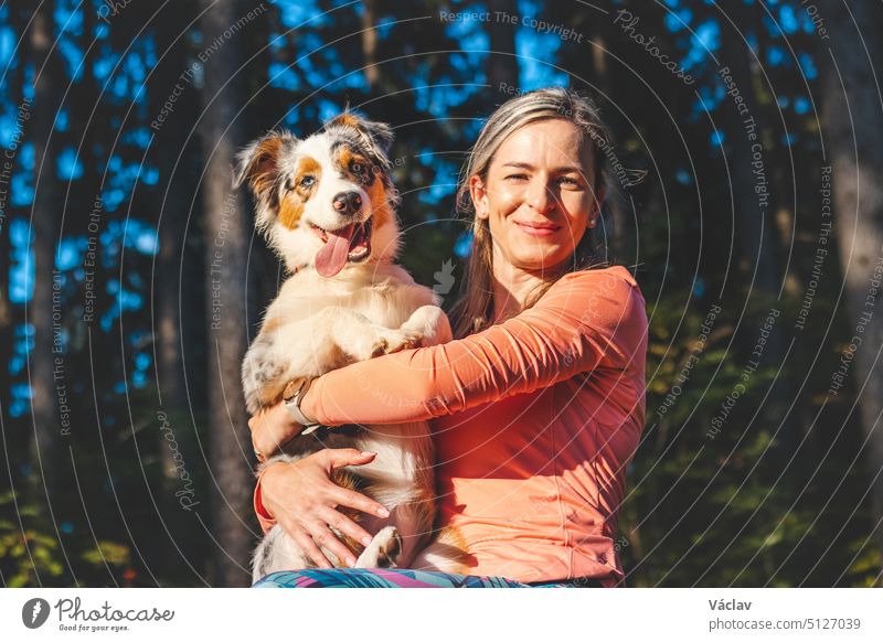 Unverblümtes Porträt einer jungen Sportlerin mit ihrem Lauf- und Wanderpartner, einem Australian Shepherd Hund. Realistisches Lächeln von Frau und Hund. Glückliches Paar. Blue Merle und Wanderer