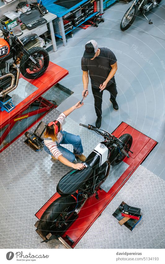 Unbekannter Mechaniker gibt einer Mitarbeiterin Werkzeug für die Reparatur eines Motorrads auf der Garaje unkenntlich Frau Partner Kollege Teamwork Paar Geben