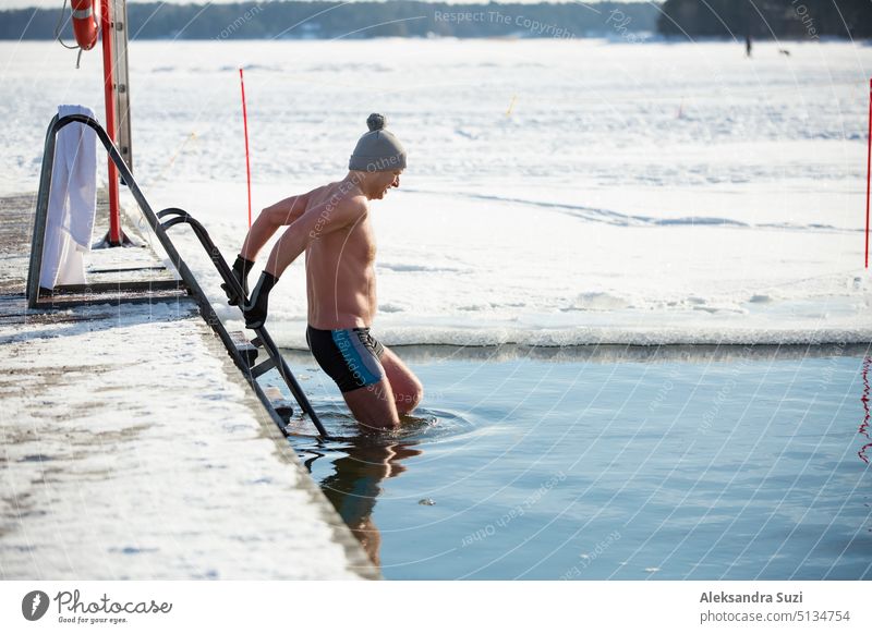 Ein Mann mit warmer Mütze und Handschuhen schwimmt in einem Eisloch und geht am Pier entlang. Winteraktivitäten in Finnland. Gesunder Lebensstil Aktivität