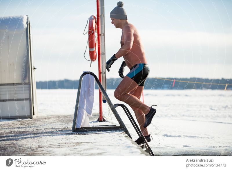 Ein Mann mit warmer Mütze und Handschuhen schwimmt in einem Eisloch und kommt aus dem Wasser. Winteraktivitäten in Finnland. Gesunder Lebensstil Aktivität