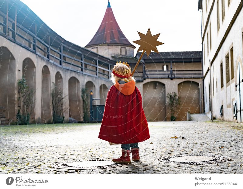 Ein kleines Kind von hinten mit rotem Cape und Krone auf einem großen Burgplatz läuft in Richtung eines Turmes und trägt einen goldenen Stern an einem Stiel vor sich her