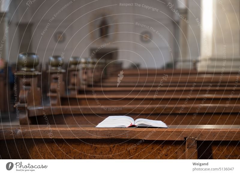 Blick über Kirchenbänke aus dunklem Holz, während im Vordergrund oder Fokus eine aufgeschlagene Bibel oder ein Gesangbuch auf der Holzbank liegt Kirchenraum