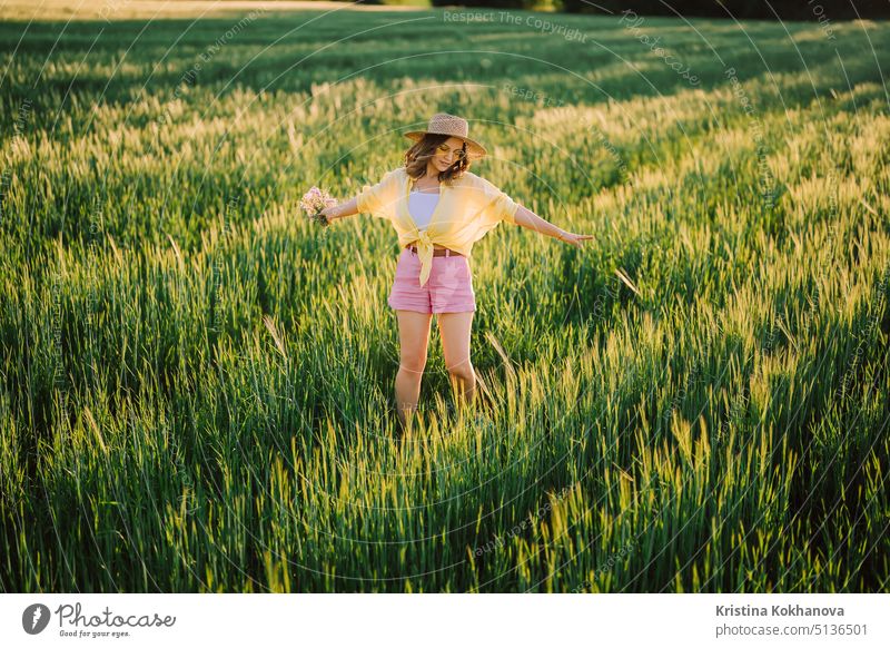 Glückliche Frau mit Strohhut tanzt in einem frischen grünen Weizenfeld. Gras Hintergrund. Erstaunliche Natur, Ackerland, wachsende Getreidepflanzen. Stilvolle Dame mit Brille.