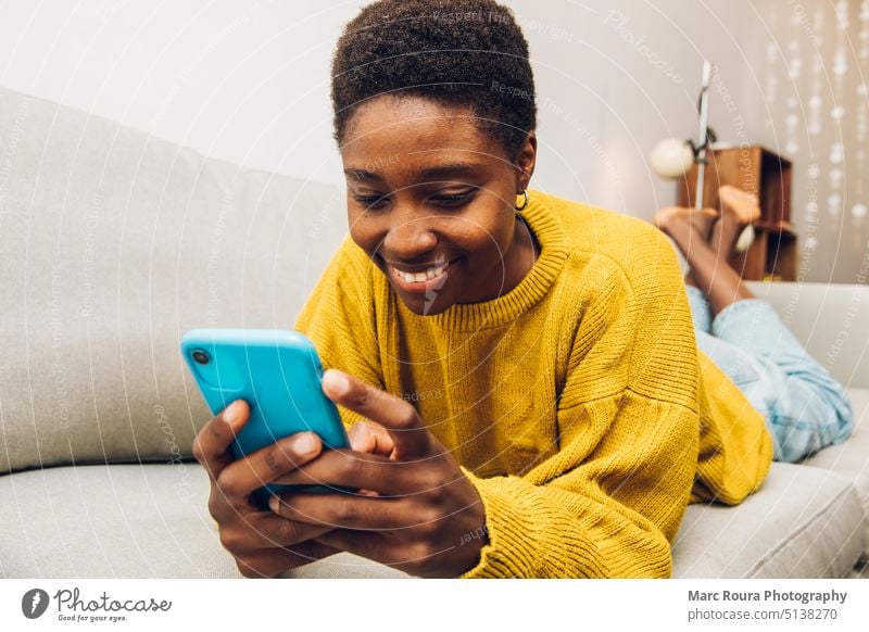 Afrikanische Frau, die zu Hause auf dem Sofa sitzend ein Mobiltelefon benutzt Erwachsener Business Anruf lässig Funktelefon Talkrunde heiter Komfort Mitteilung
