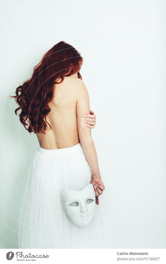 Eine Frau mit langen roten Haaren steht mit dem Rücken zur Kamera. Sie ist oberkörperfrei, trägt einen weißen Tüllrock und hat eine weiße Maske in der Hand.