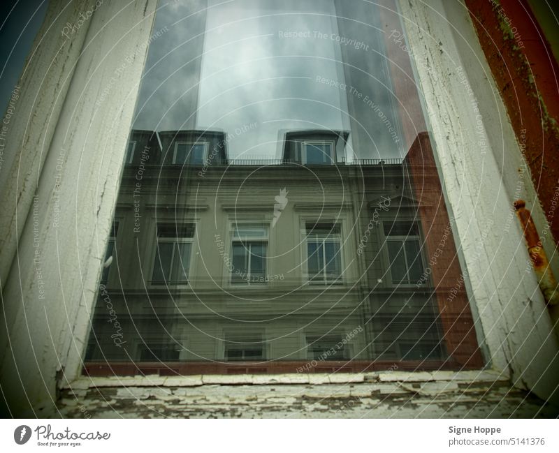 In einer Fensterscheibe mit brüchigem Holzrahmen spiegelt sich an einem düsteren Tag das gegenüberliegende Altstadthaus. Spiegelung fenster fensterrahmen holz